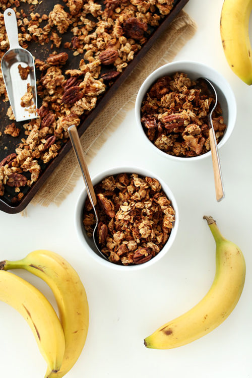 Baking sheet and bowls of Banana Bread Granola alongside fresh bananas