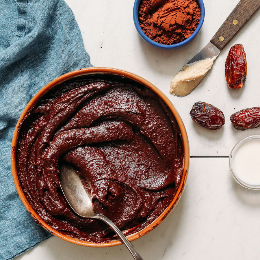 Bowl of oil-free chocolate frosting next to cocoa powder, cashew butter, dates, and dairy-free milk
