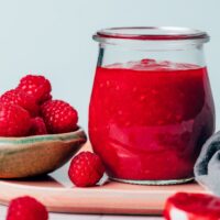 Close up photo of a jar of raspberry compote next to a bowl of fresh raspberries