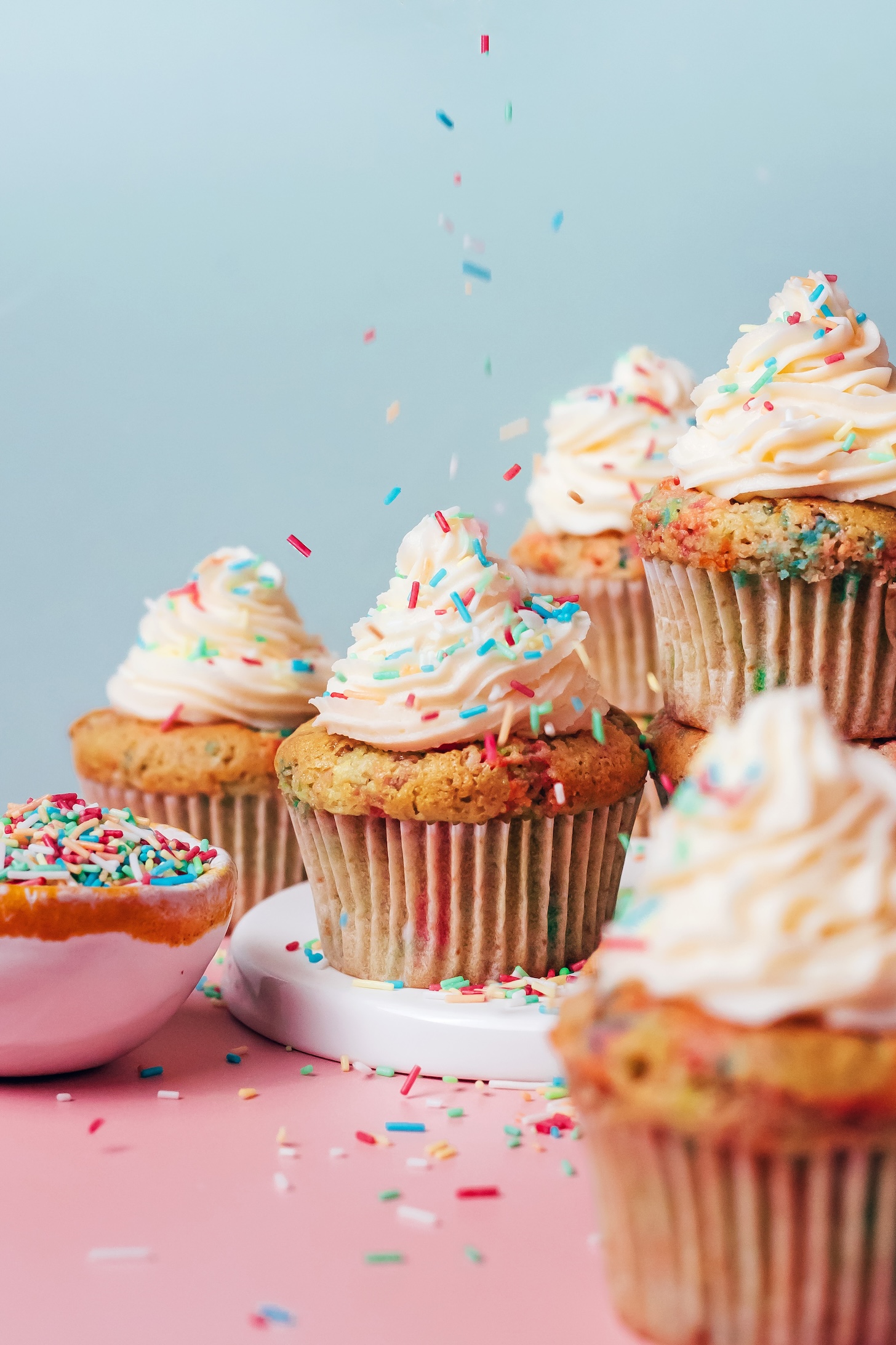 Sprinkling rainbow sprinkles over a batch of frosted funfetti cupcakes