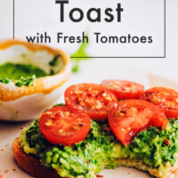 Slice of vegan pesto avocado toast with fresh tomatoes and a bowl of vegan pesto behind