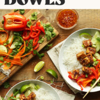 Two servings of our veggie noodle bowls with almond butter tofu next to lots of fresh veggies and a sauce